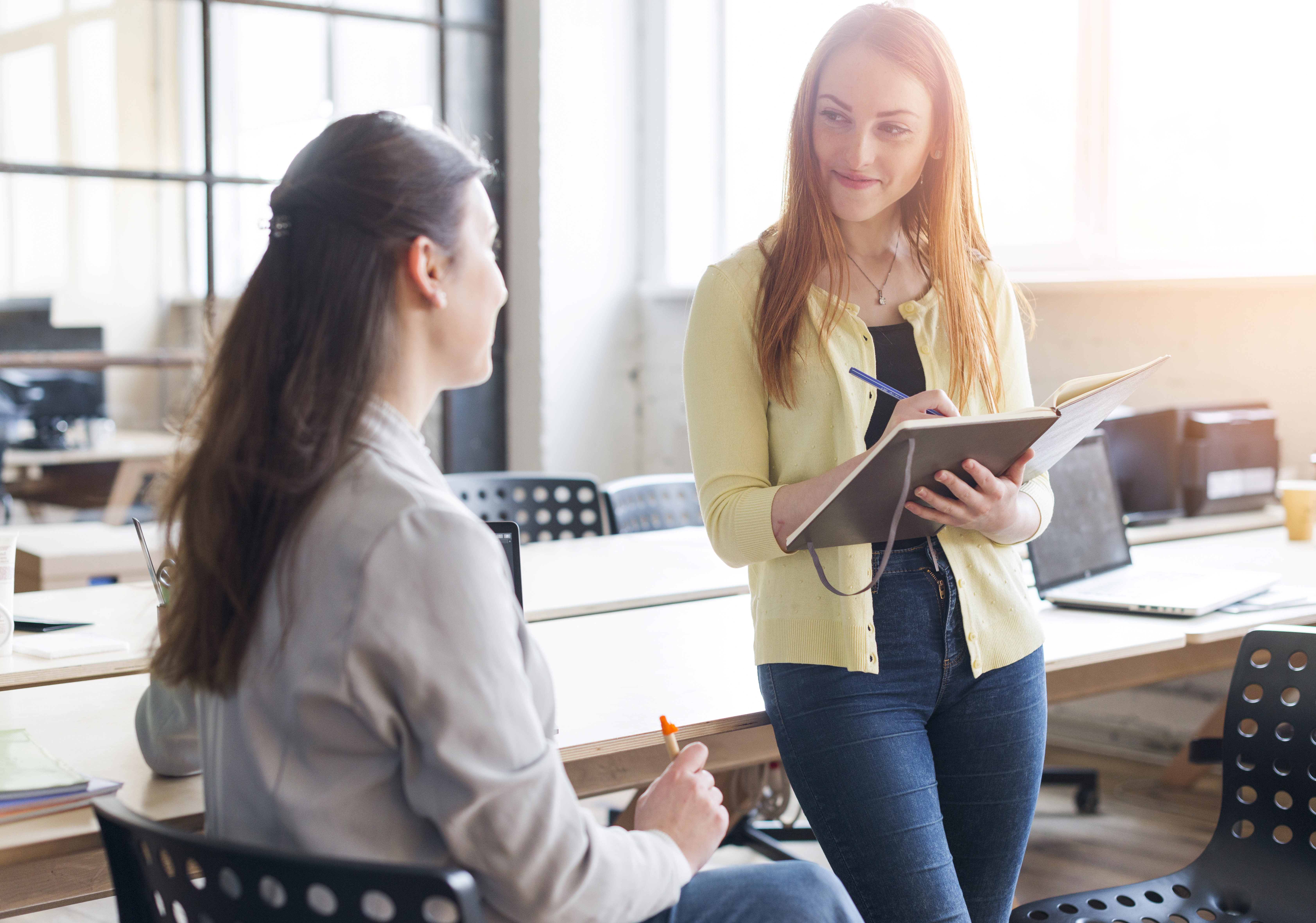 smiling-woman-holding-diary-pen-looking-her-colleague-office